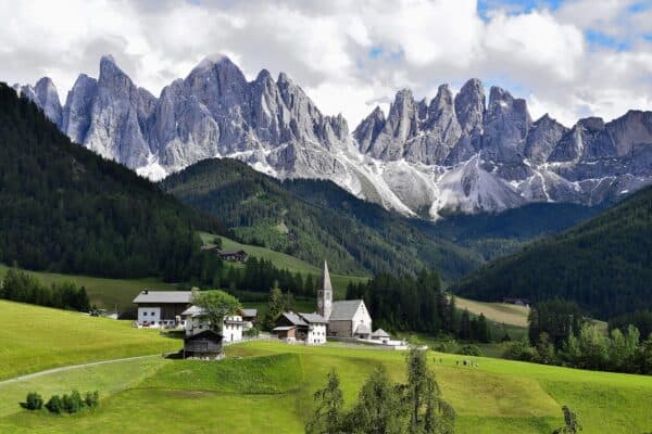 Majestic Dolomites mountain range in Italy under a clear blue sky with scattered clouds, showcasing alpine peaks, lush green valleys, and a panoramic landscape ideal for hiking.