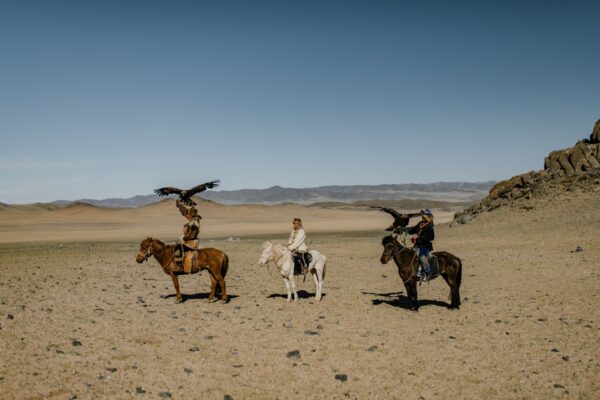 Nomadic Mongolian herders on horseback traverse vast golden desert dunes, with majestic golden eagles perched alertly on their outstretched arms, wings poised for flight. 